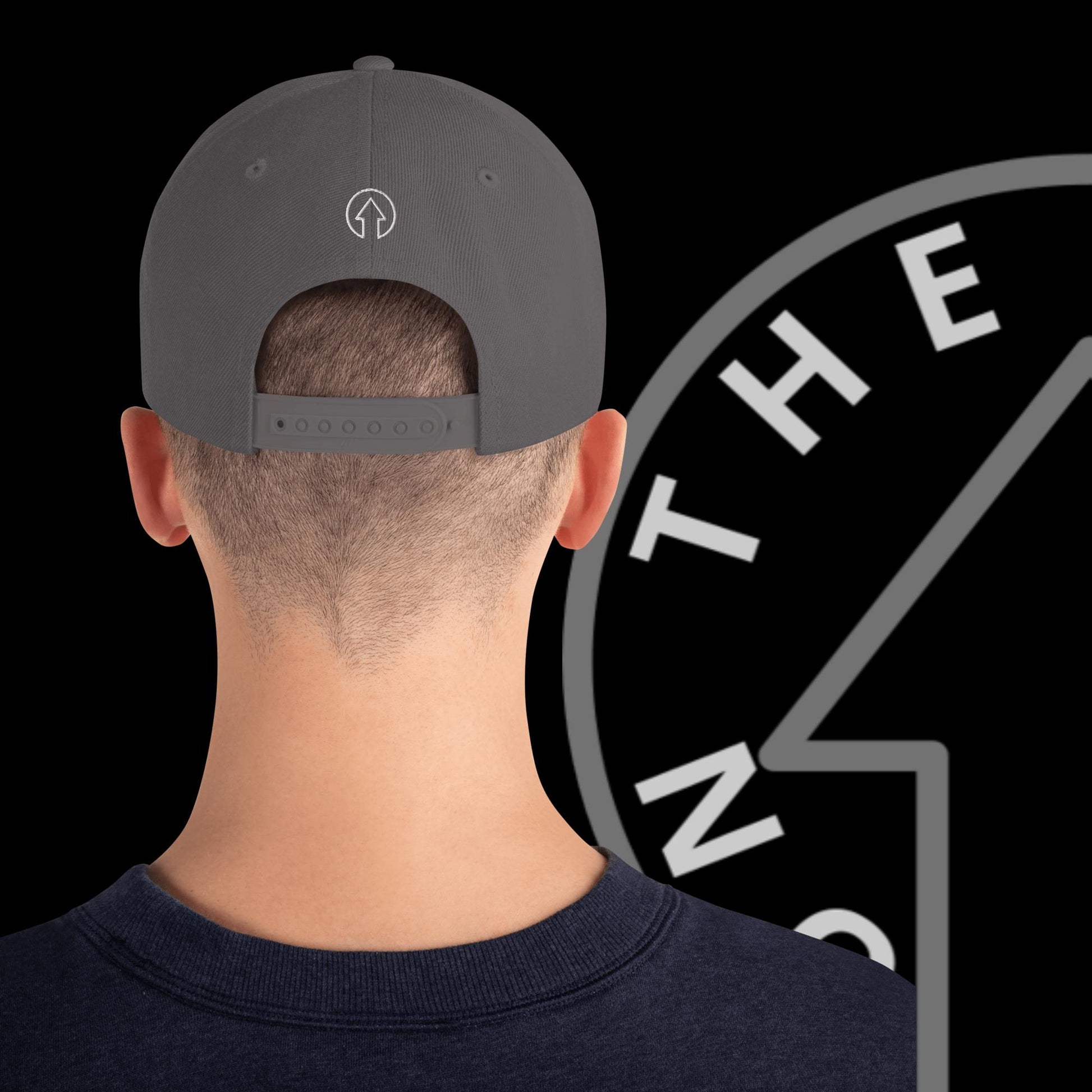 Rear view of a man wearing a gray Signature On The Way Up Logo Snap Back backwards, showing a visible hairline on his neatly trimmed head against a dark background with partial white letters.