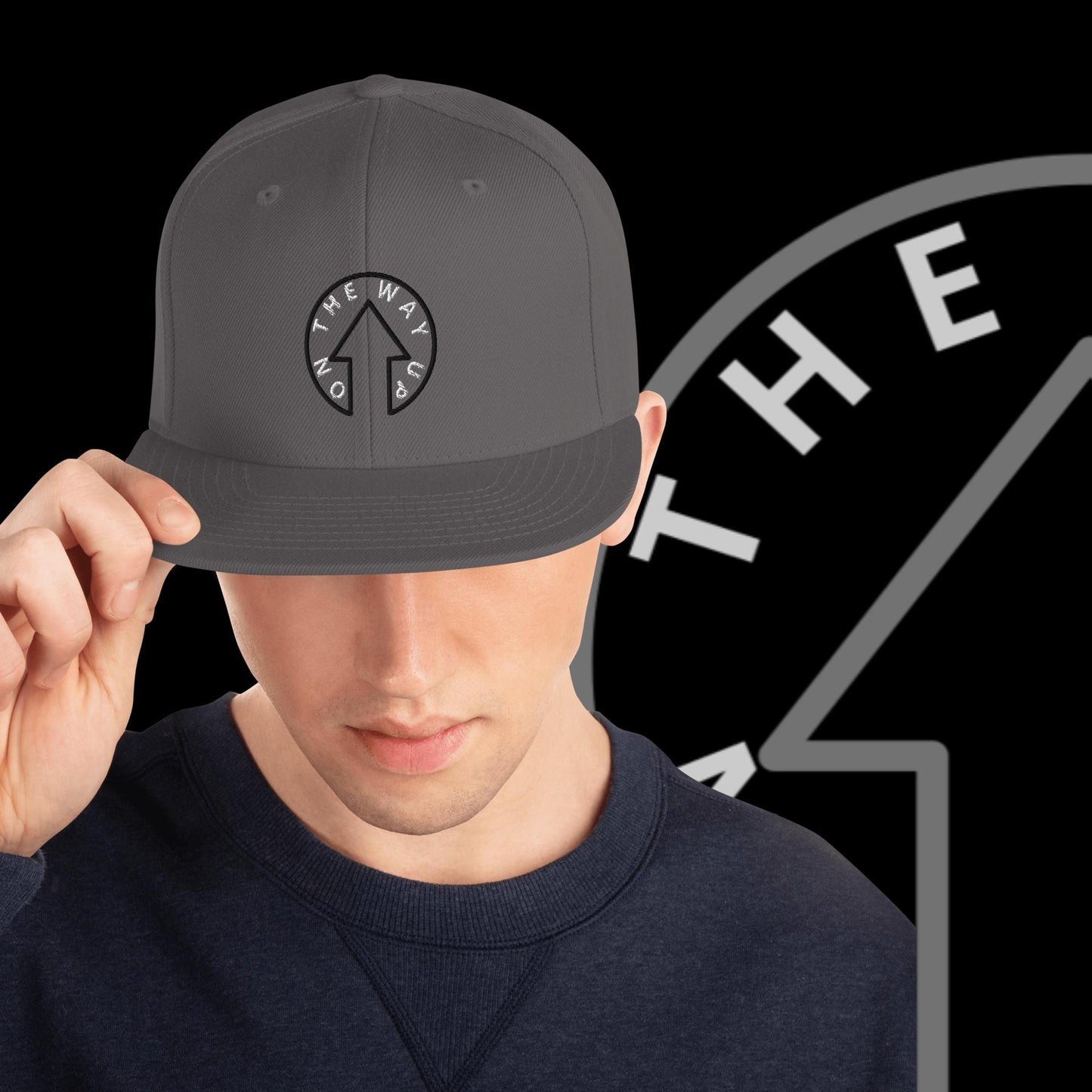 A young man tilting a gray baseball cap from On The Way Up Apparel with the Signature On The Way Up Logo Snap Back on his head, set against a black background with partial "THE" lettering.