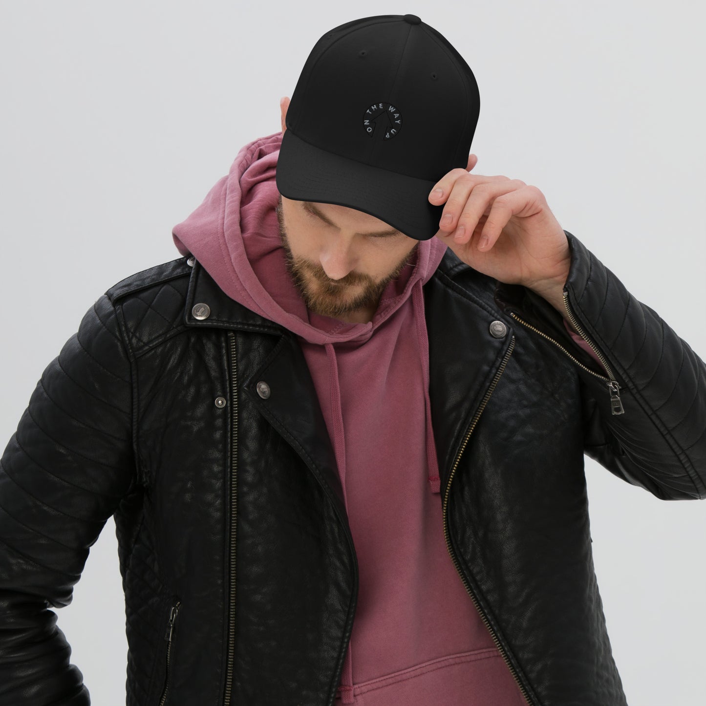 A man in a stylish layered outfit adjusting his Stretch Fit On The Way Up Signature Cap. He wears a premium quality black leather jacket over a rose-colored hooded sweatshirt, set against a plain background from On The Way Up Apparel.