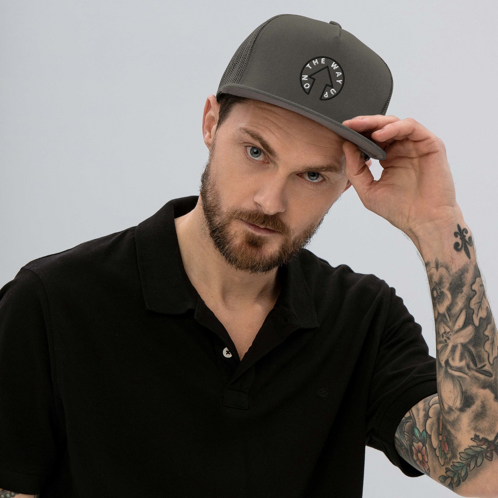 A man with a beard, wearing a premium quality black polo shirt and an On The Way Up Signature Trucker Cap, adjusts his hat while looking intently at the camera. His arm, showing a sleeve tattoo, slightly lifts the cap's brim.