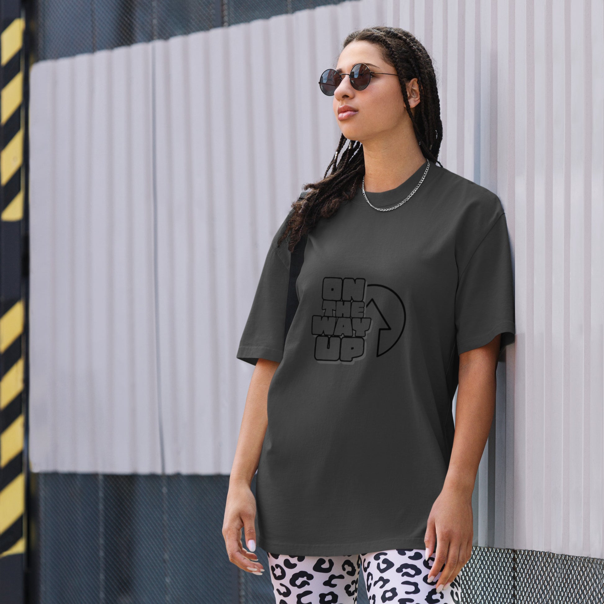 A person with long hair and sunglasses stands against a metal wall with a yellow and black diagonal striped pattern on the edges. They wear an oversized grey "Oversized faded Large print t-shirt" from On The Way Up Apparel, paired perfectly with white leopard print leggings.