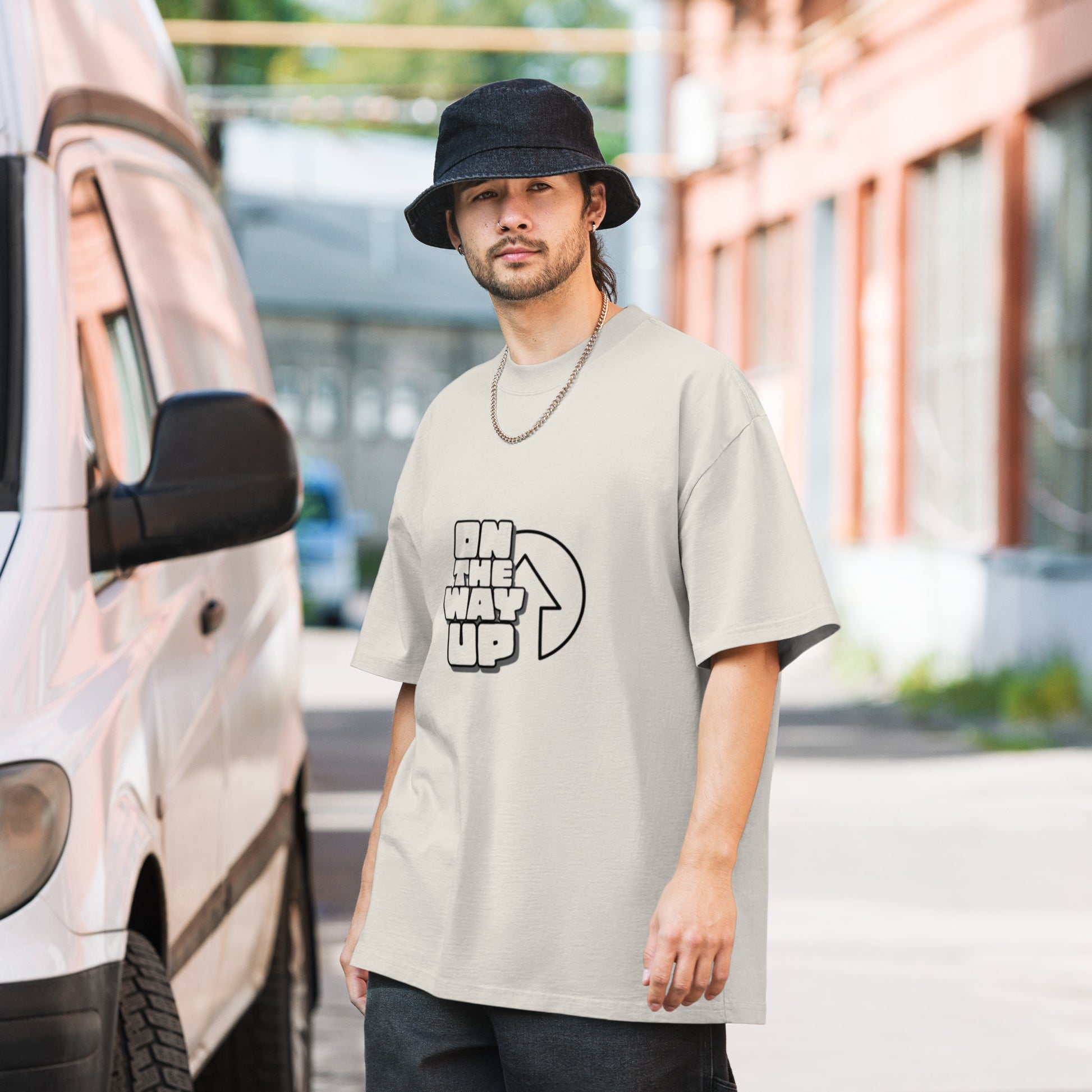 A man stands next to a white van on a sunny street, wearing a black bucket hat and a beige oversized faded Large print t-shirt made of 100% carded cotton from On The Way Up Apparel with the text "ON THE WAY UP" and an upward arrow graphic. The background features buildings with red and brown tones.