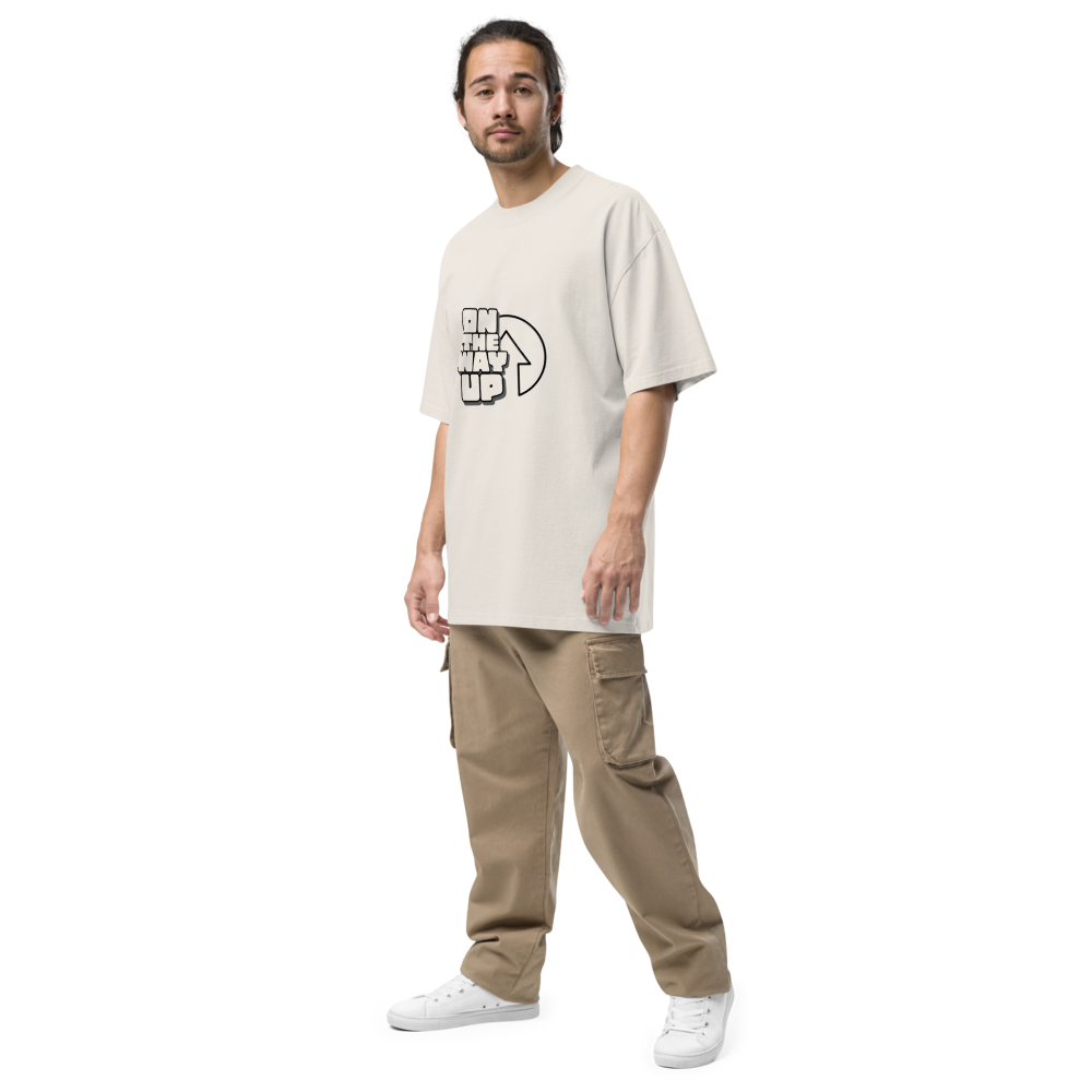 A person with medium-length hair, wearing an oversized faded Large Print t-shirt from On The Way Up Apparel and light brown cargo pants, stands against a plain black background. They are also wearing white sneakers and looking towards the camera.