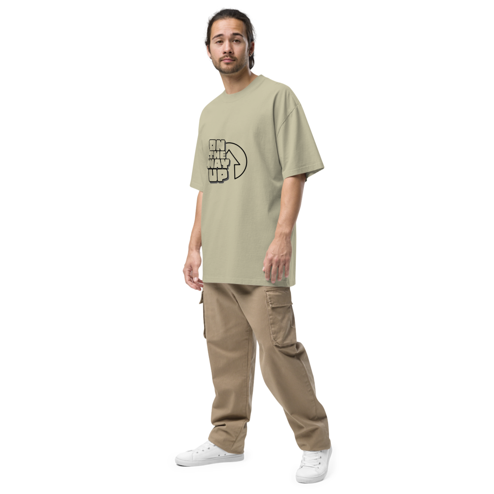 A person with shoulder-length hair stands facing forward in a green oversized faded Large Print t-shirt from On The Way Up Apparel made from 100% cotton, beige cargo pants, and white sneakers. They have a relaxed posture with one hand by their side and the other slightly out. The background is plain white.