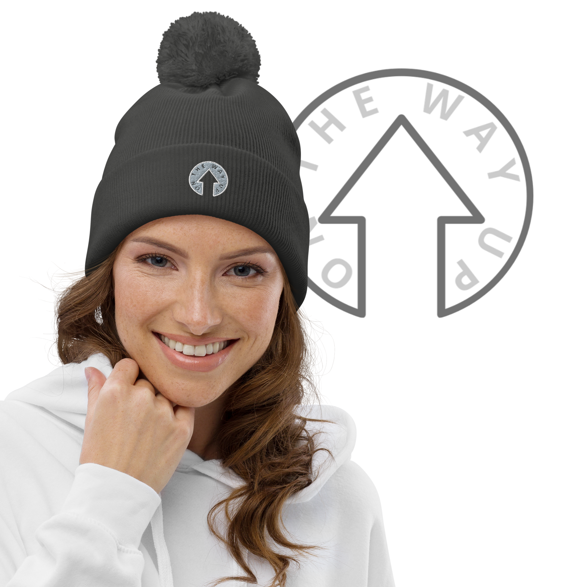 A smiling woman with light skin wearing a white hoodie and the On The Way Up Logo Pom pom beanie, standing against a black background with a partial On The Way Up Apparel circular logo.