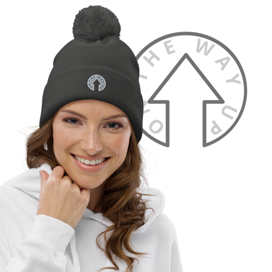 A smiling woman with light skin wearing a white hoodie and the On The Way Up Logo Pom pom beanie, standing against a black background with a partial On The Way Up Apparel circular logo.