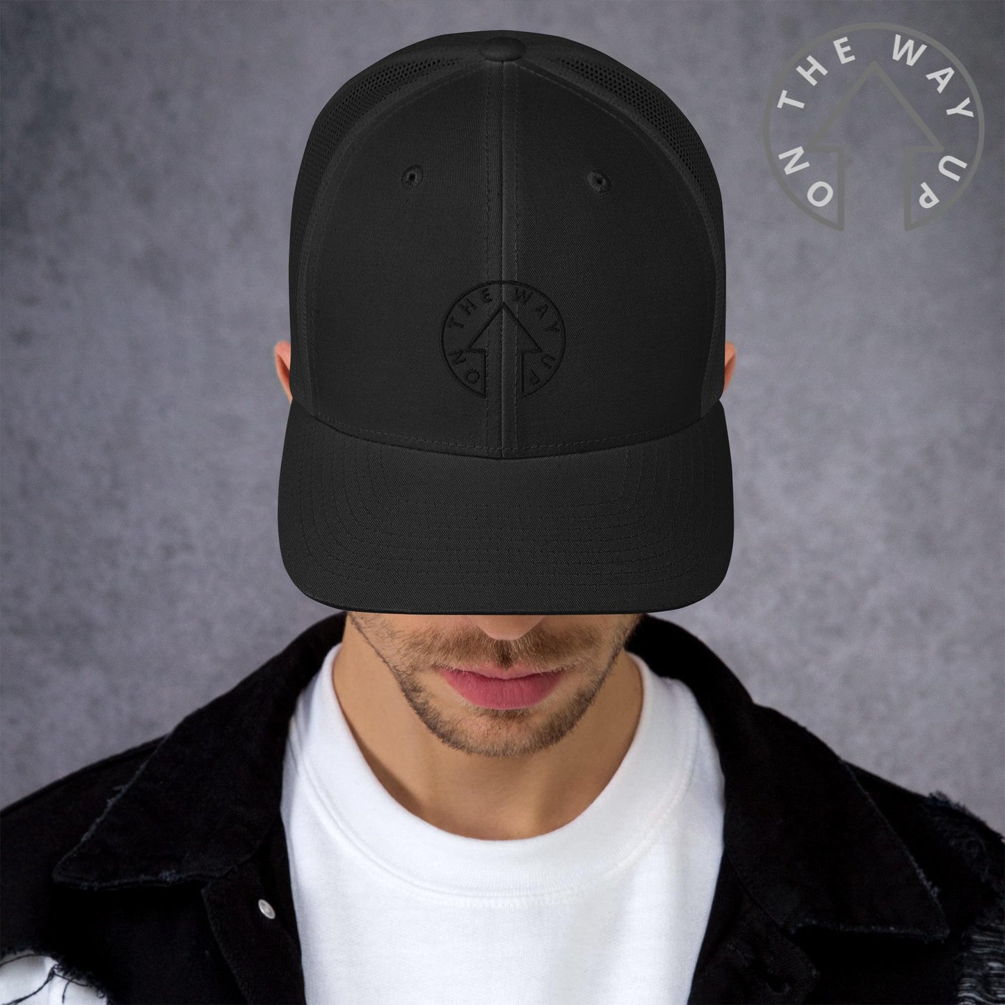 A young man wearing an On The Way Up Apparel black baseball cap and a black jacket, only his lower face visible, stands against a textured gray background. The cap features a circular logo with the words "Manchester Premium Quality.