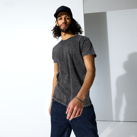 A confident man with long curly hair and a beard, wearing a grey Signature Denim Tee by On The Way Up Apparel, jeans, and a black baseball cap branded with "Manchester," is standing in a studio with a striking shadow on the wall.