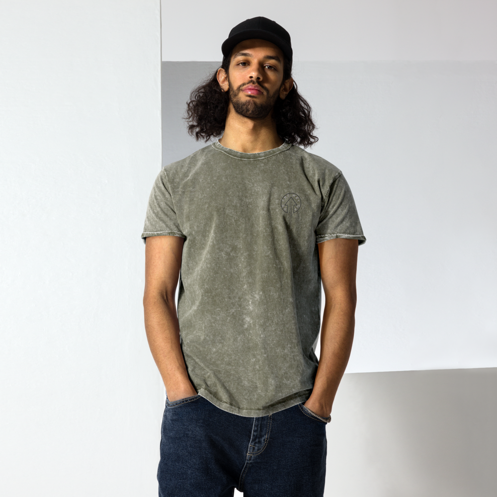 A man with shoulder-length curly hair, donned in a Signature Denim Tee from On The Way Up Apparel and dark jeans, stands confidently with his hands in his pockets against a white background.