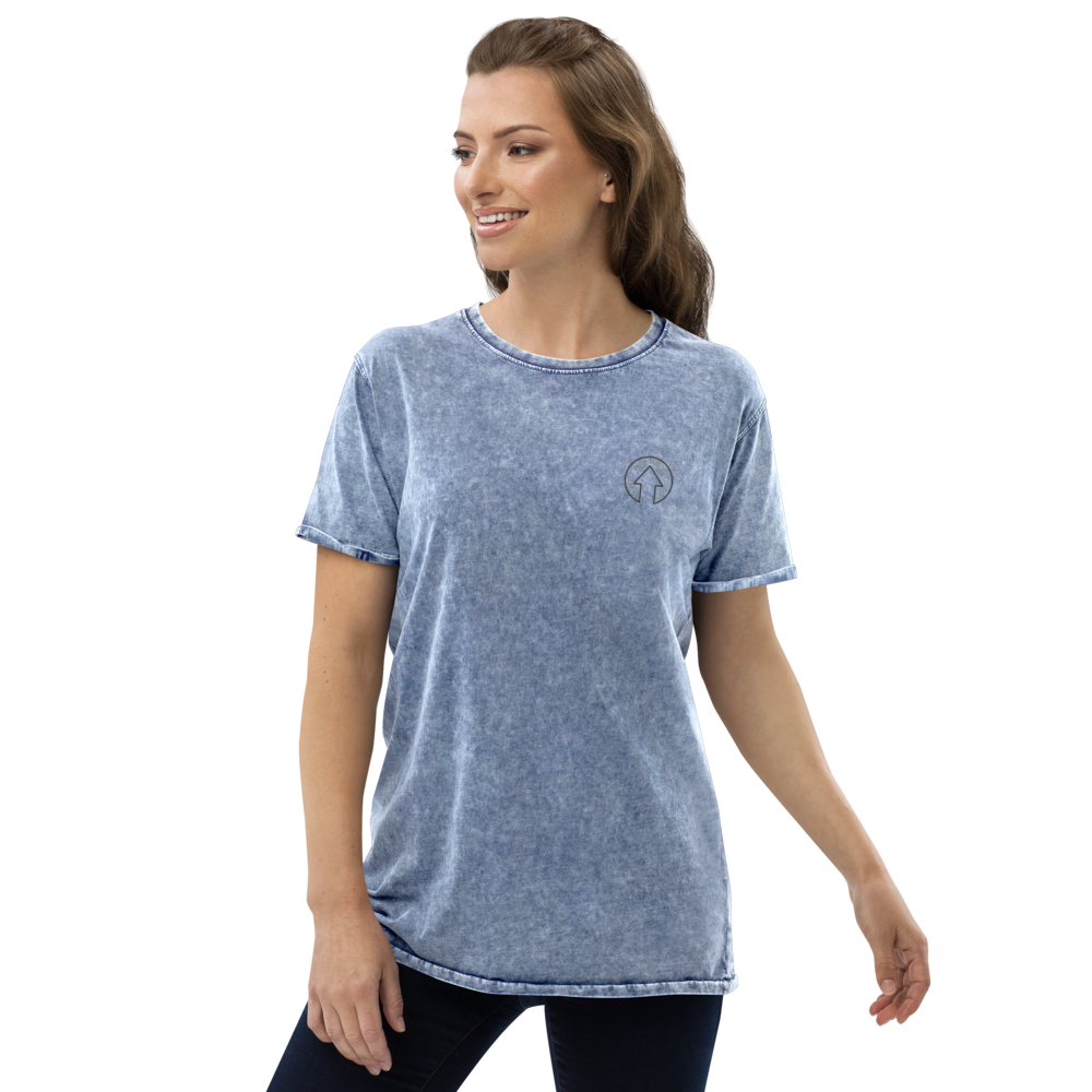 A woman wearing an On The Way Up Apparel Signature Denim Tee, a premium quality blue acid-wash t-shirt with a small logo on the left chest, smiling and posing against a transparent background.