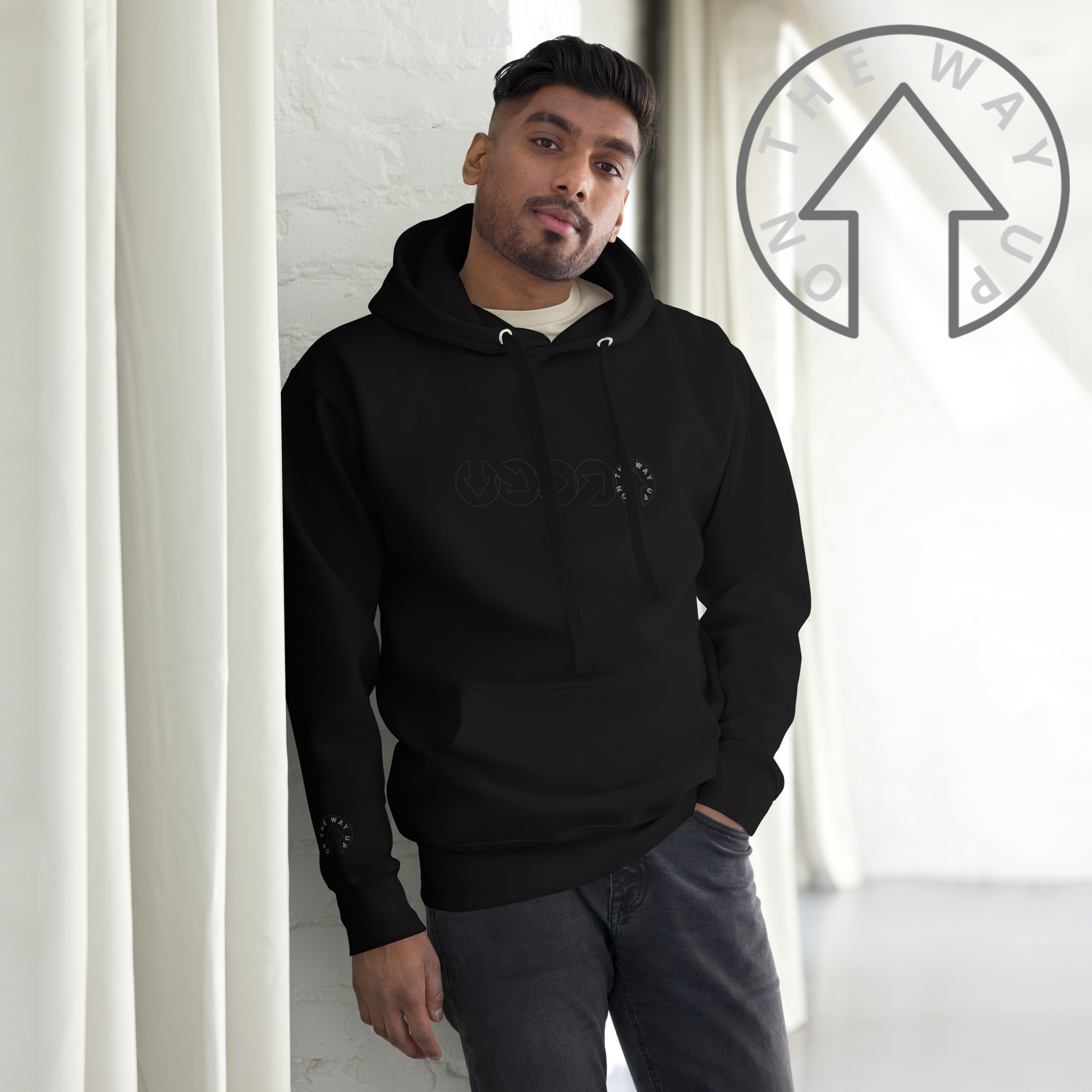 A young man in an On The Way Up Progress Hoodie from On The Way Up Apparel stands confidently in a bright room with white walls, gazing thoughtfully towards the camera. His attire screams premium quality, capturing the essence of modern urban fashion.