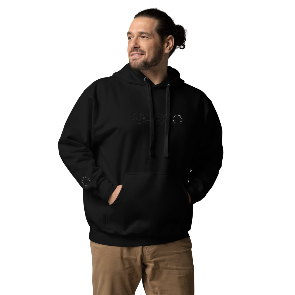 A man in an On The Way Up Apparel premium quality black hoodie and beige pants, standing with hands in pockets and smiling slightly, looks to his right. The hoodie features a white logo on the chest.