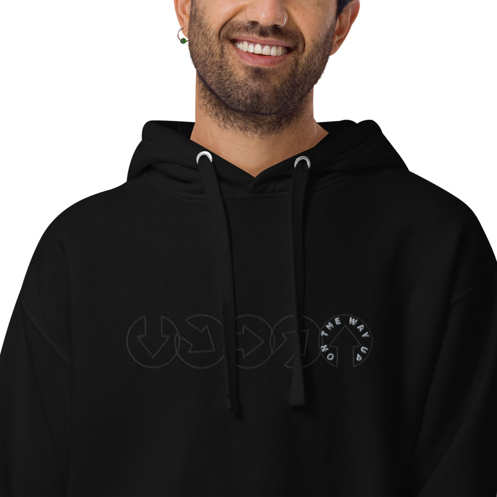 A smiling man wearing a black On The Way Up Progress Hoodie with a subtle circular "Manchester" logo design on the front. His expression is friendly and he has a neatly trimmed beard.