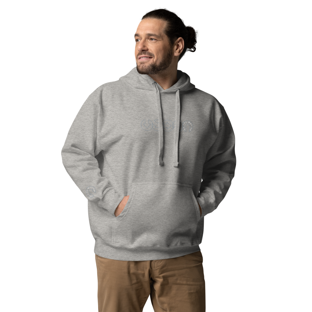 A man wears the On The Way Up Progress Hoodie in Grey and khaki pants, hands in pockets, smiling slightly as he looks to his right in front of a black background.