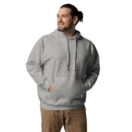 A man wears the On The Way Up Progress Hoodie in Grey and khaki pants, hands in pockets, smiling slightly as he looks to his right in front of a black background.