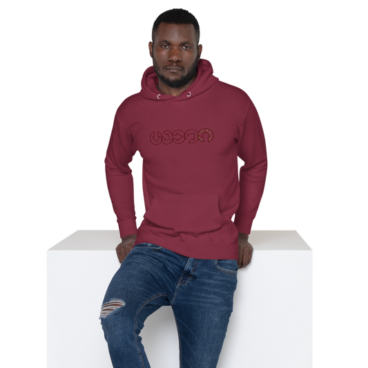 A man in a maroon On The Way Up Progress Hoodie - Cherry and distressed blue jeans sitting on a white block, looking directly at the camera. The hoodie has "Manchester" in a unique text design on the chest.