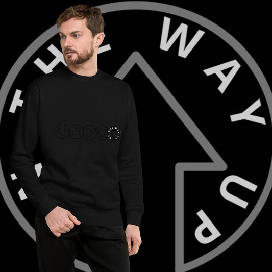 A man with a beard modeling an On The Way Up Apparel On The Way Up Progress black round neck jumper with a subtle graphic design, standing against a background featuring a round, stylized "ONE WAY" sign.