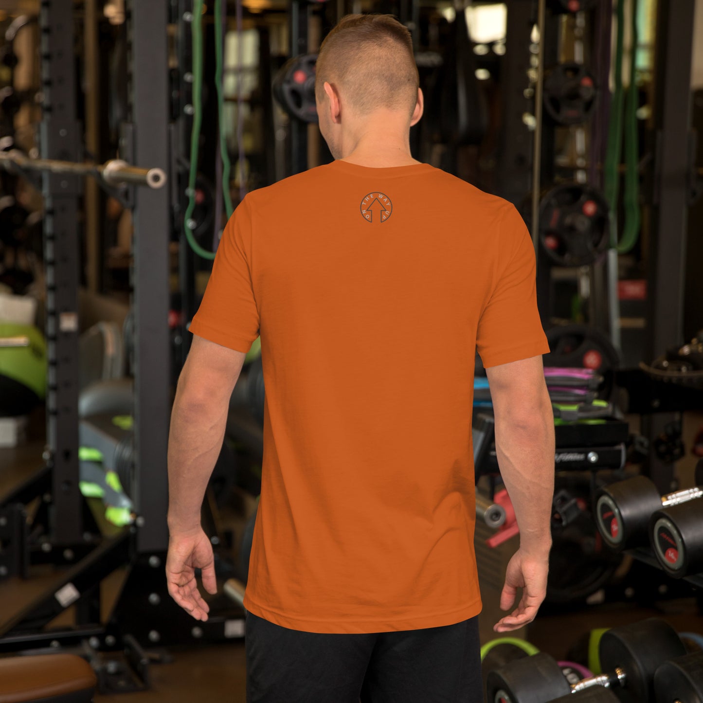 A man wearing an On The Way Up Apparel BE BOLD - On The Way Up Embroidered Logo T-shirt in autumn orange stands in a gym filled with various exercise equipment.