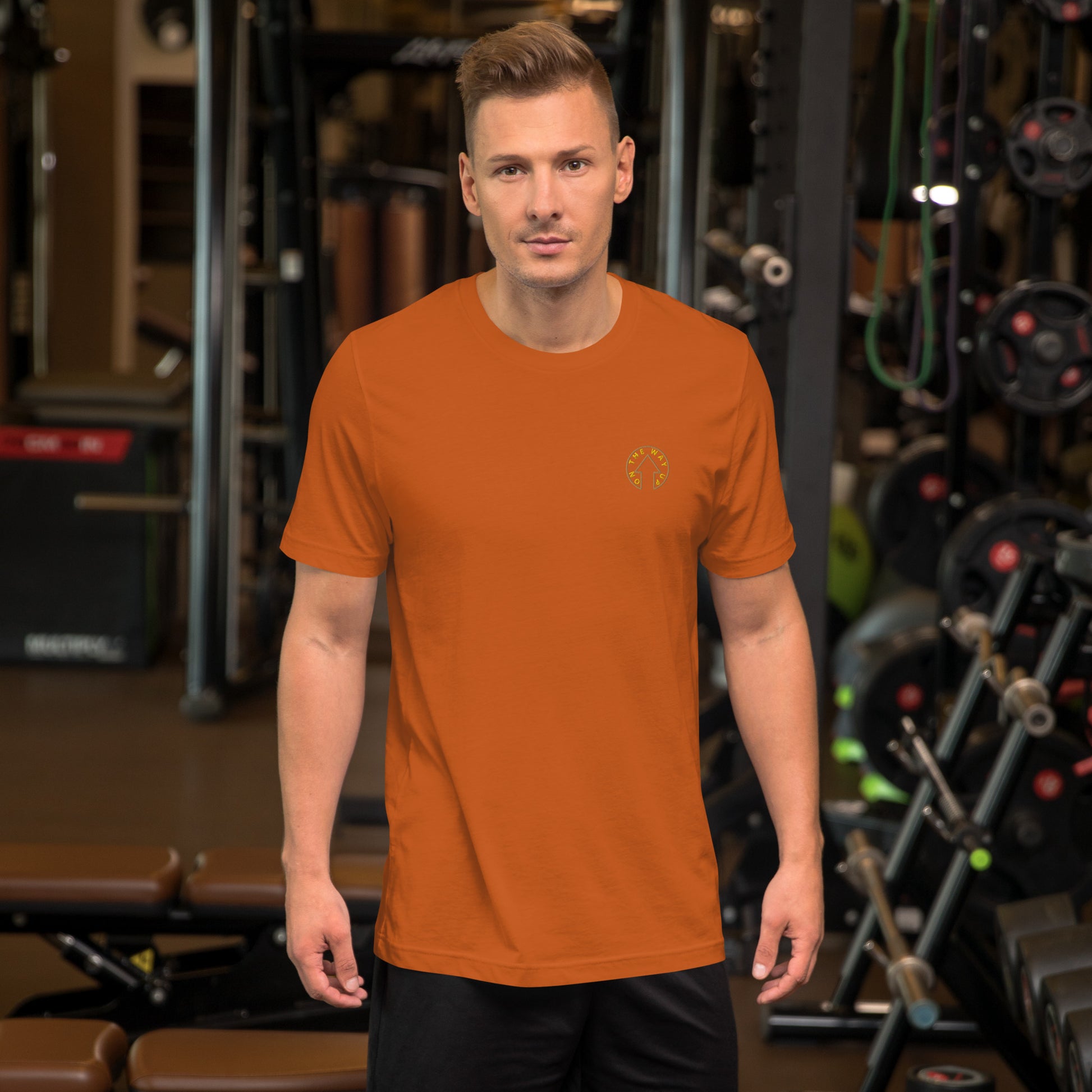 A man in an BE BOLD - On The Way Up Embroidered Logo T-shirt - AUTUMN stands confidently in a Manchester gym, surrounded by weightlifting equipment. His expression is focused and determined.