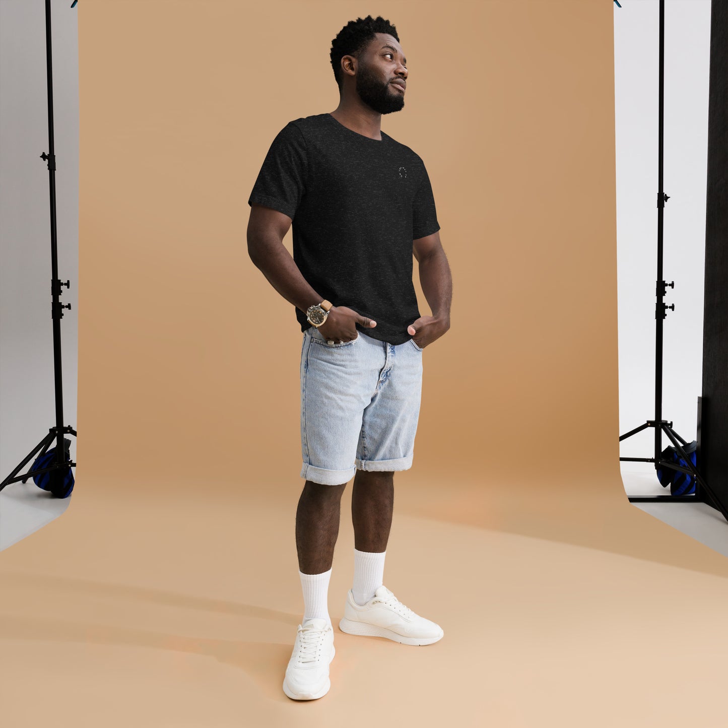 A man in a casual outfit stands in a photography studio, glancing off to the side. He wears a black Signature On The Way Up Embroidered Logo T-shirt from On The Way Up Apparel, denim shorts, and white sneakers, with a watch on his wrist.