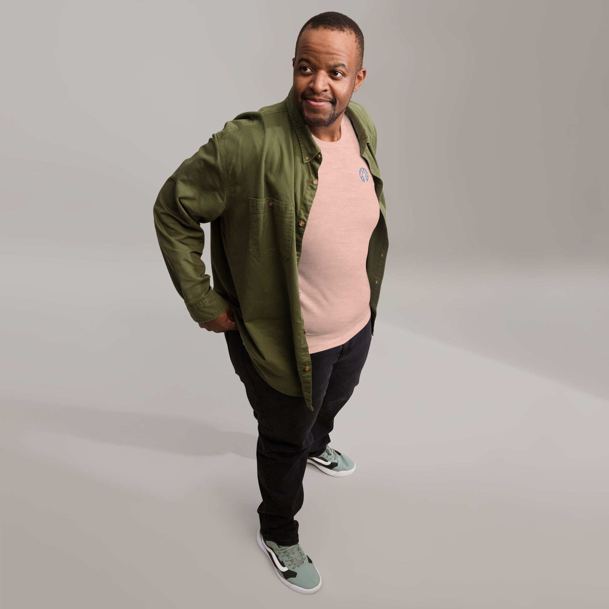 A man in casual attire stands confidently with his hand on his hip, smiling at the camera. He wears a premium quality green jacket, BE BOLD - On The Way Up Embroidered Logo T-shirt in peach, black pants, and teal sneakers against a light gray background by On The Way Up Apparel.