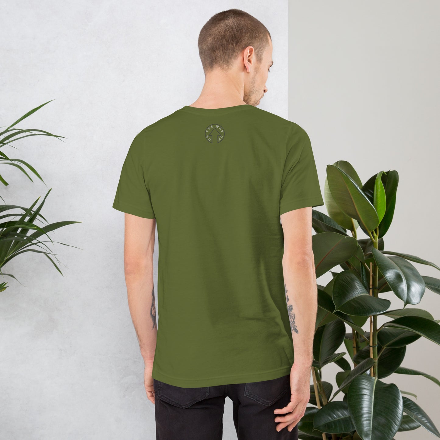 A man wearing an On The Way Up Apparel BE BOLD - On The Way Up Embroidered Logo T-shirt in OLIVE, featuring a small logo at the upper back, seen from behind, standing beside indoor plants against a gray and white background.