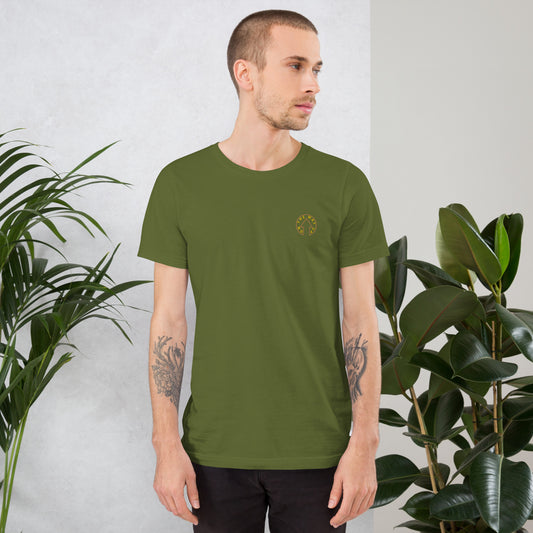 A man with tattoos on his arms, wearing an On The Way Up Apparel BE BOLD - Embroidered Logo T-shirt in OLIVE, stands confidently in an indoor setting with a potted plant in the background.