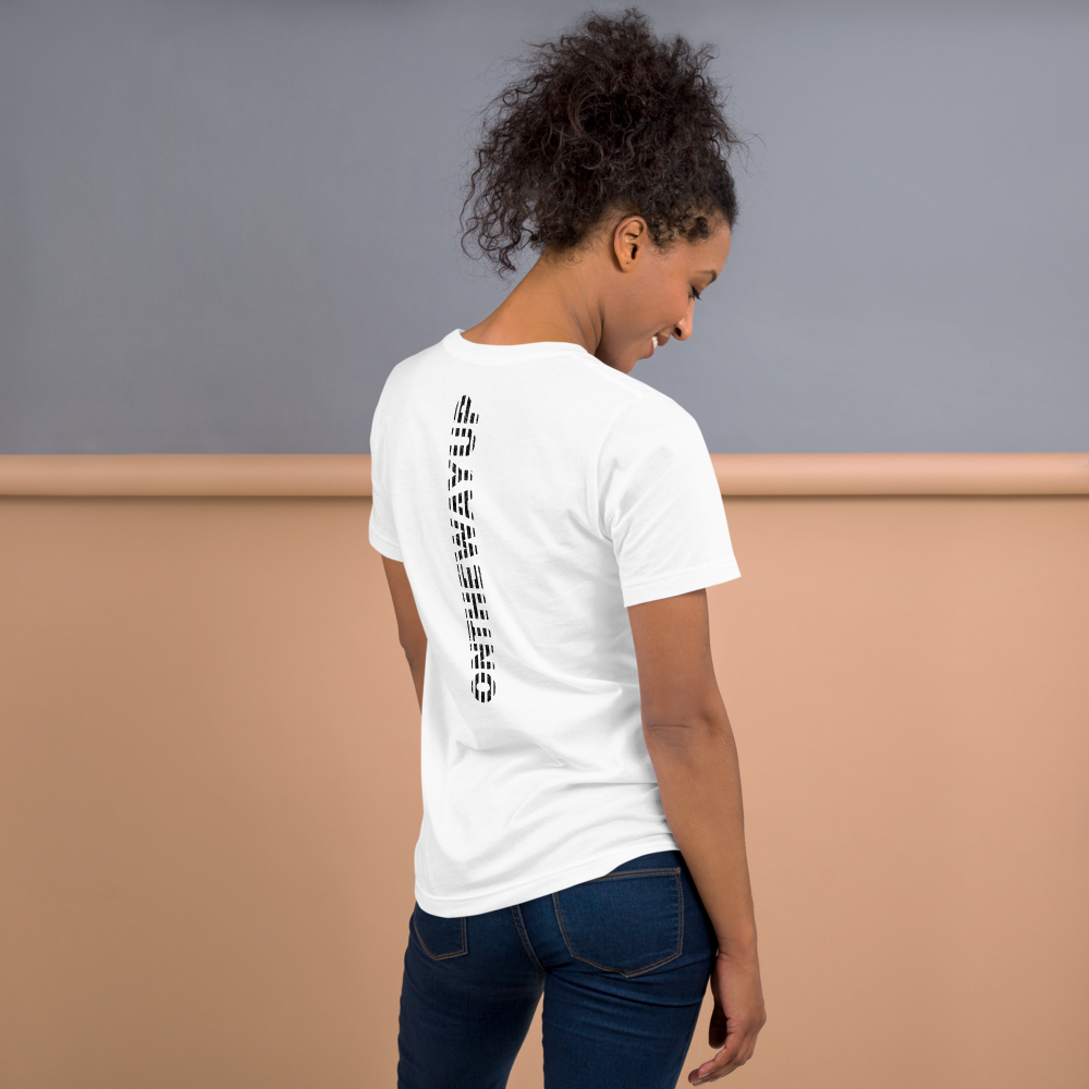 A person with curly hair is standing with their back to the camera, wearing the Scribble Logo T-shirt by On The Way Up Apparel, paired with blue jeans. The lightweight, white tee is crafted from combed and ring-spun cotton and features a vertical black design down the back. They are positioned against a two-tone wall in gray and peach colors.