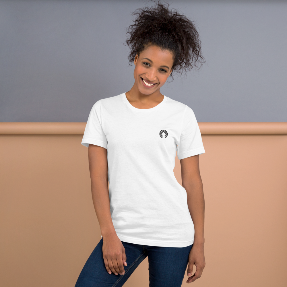 A person with curly hair smiling, wearing a Scribble Logo T-shirt by On The Way Up Apparel and blue jeans. The white t-shirt, made of lightweight cotton and pre-shrunk fabric, features a small black circular logo on the left side. It pairs perfectly with the two-tone background: the top half in gray and the bottom half in soft peach.