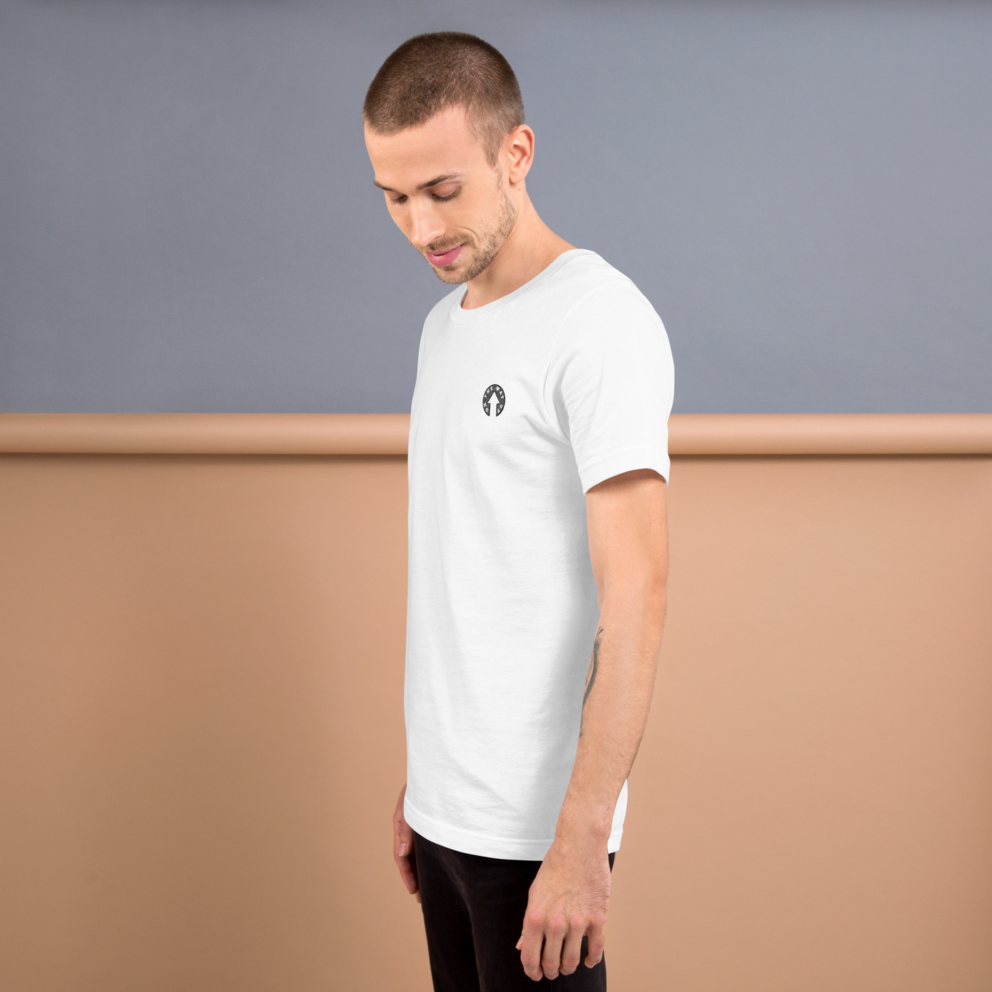 A man with short hair and a trimmed beard is standing in a room, facing slightly to the side. He is wearing a plain white Scribble Logo T-shirt from On The Way Up Apparel and black pants. The background features a gradient of blue and beige walls.