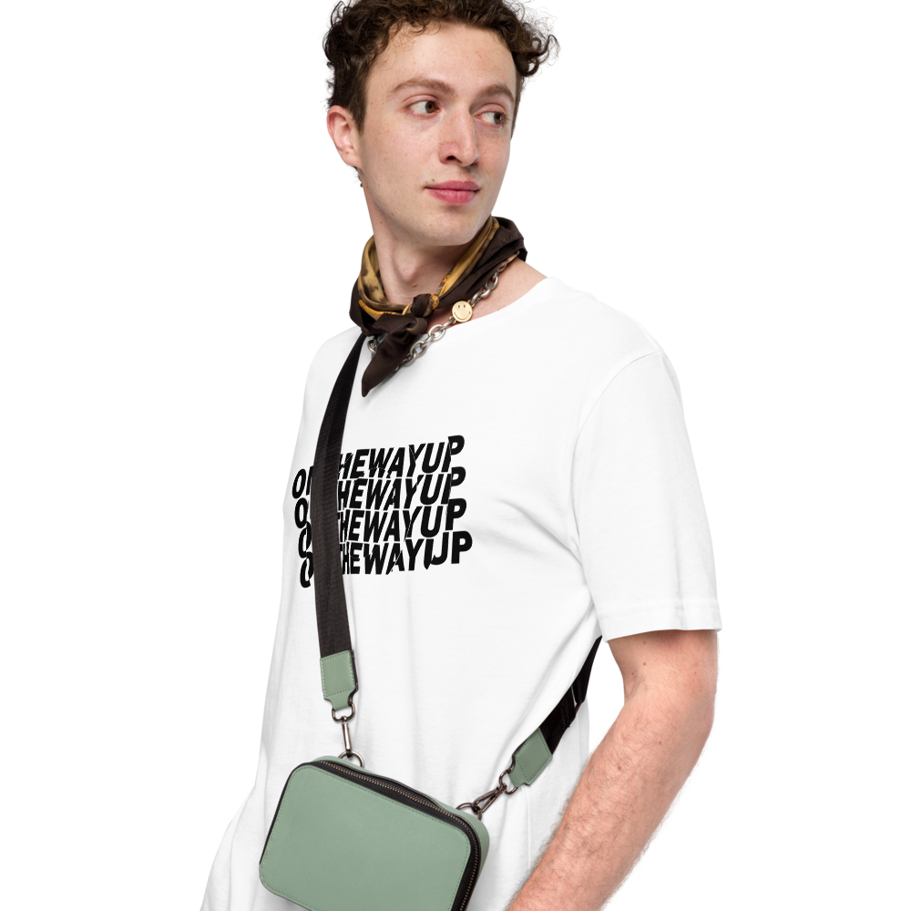 A young man with curly hair is wearing the Circular Rotation Slogan Tee from On The Way Up Apparel, a white T-shirt made of super soft, combed and ring-spun cotton featuring the repetitive "THE WAY UP" text. He has a brown and yellow scarf around his neck and a green crossbody bag. Looking to his left with a faint smile, he stands against a plain white background.