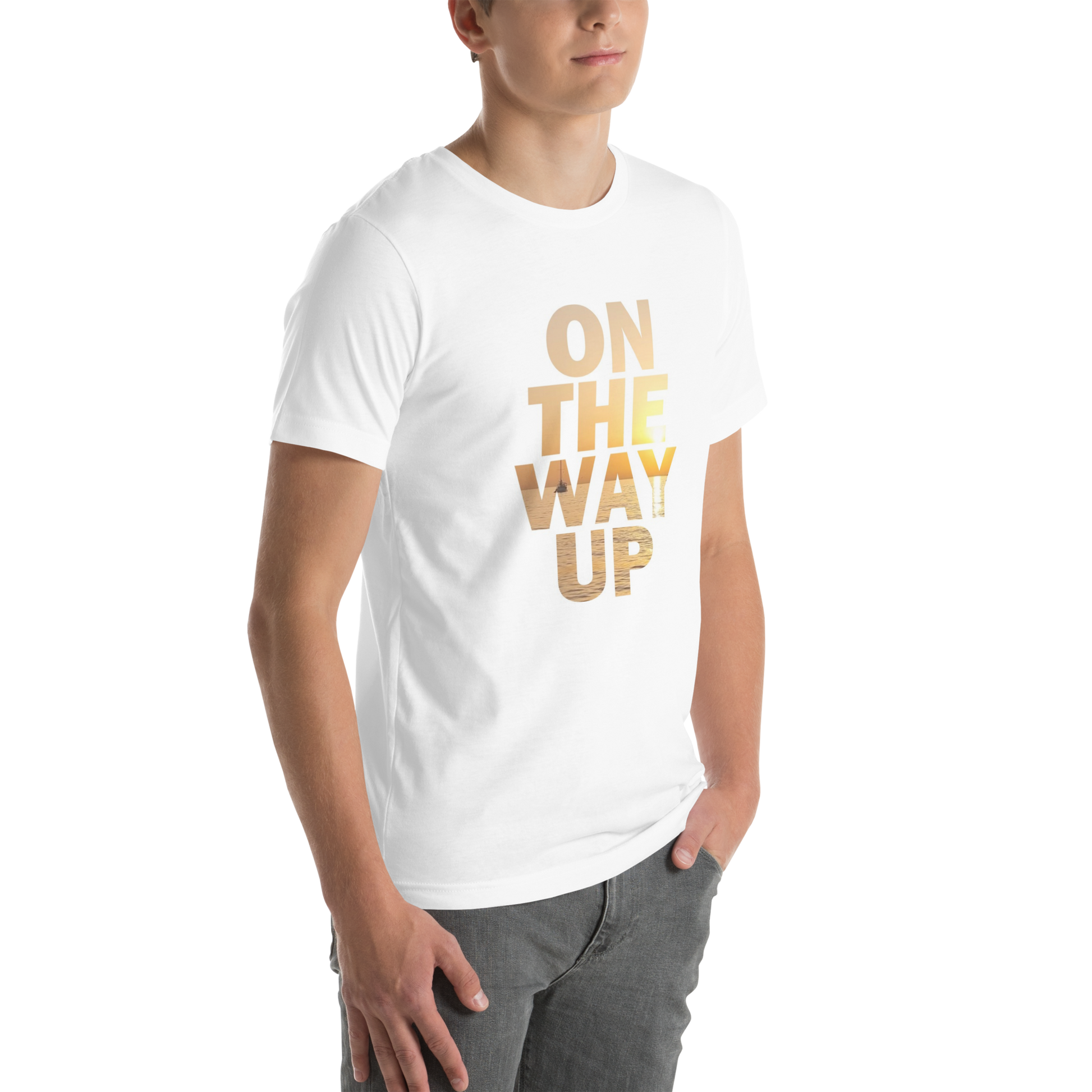 A person stands against a black background wearing the Mambo Beach Sunset Tee from On The Way Up Apparel. The comfy and cool white T-shirt features the text "ON THE WAY UP!" printed in gradient shades of gold and yellow. Made from lightweight cotton, they have one hand in their pocket and the other arm relaxed by their side, partially facing the camera.