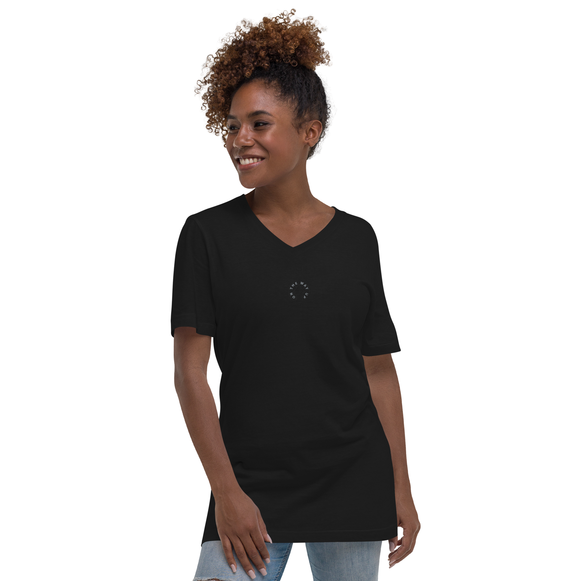 A joyful young Black woman with curly hair wearing an On The Way Up Apparel Signature Short Sleeve V-Neck T-Shirt in black and jeans, standing and looking to her left with a big smile.