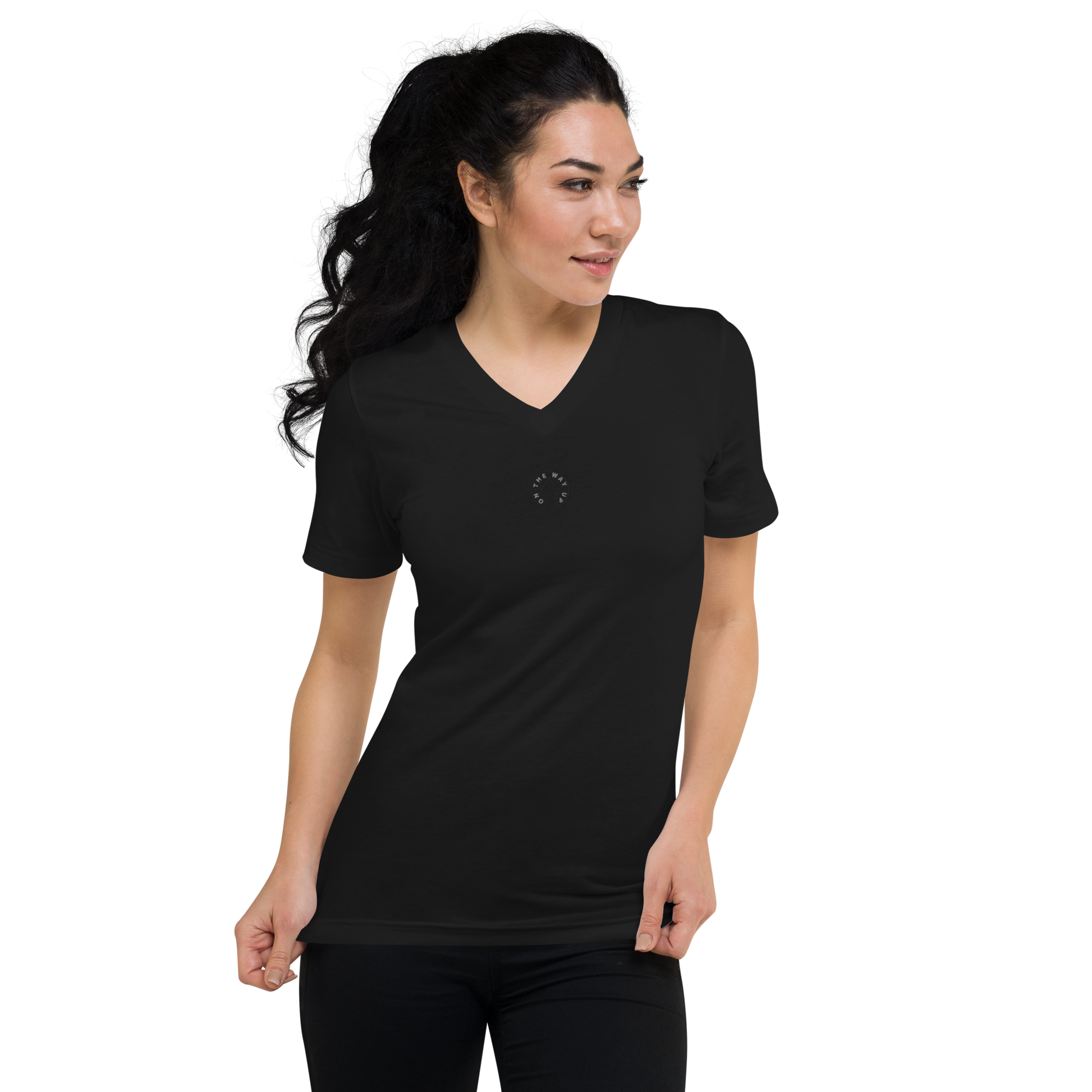 A woman in a On The Way Up Apparel Signature Short Sleeve V-Neck T-shirt and black pants standing against a dark background, looking to her left with her hair blowing behind her.